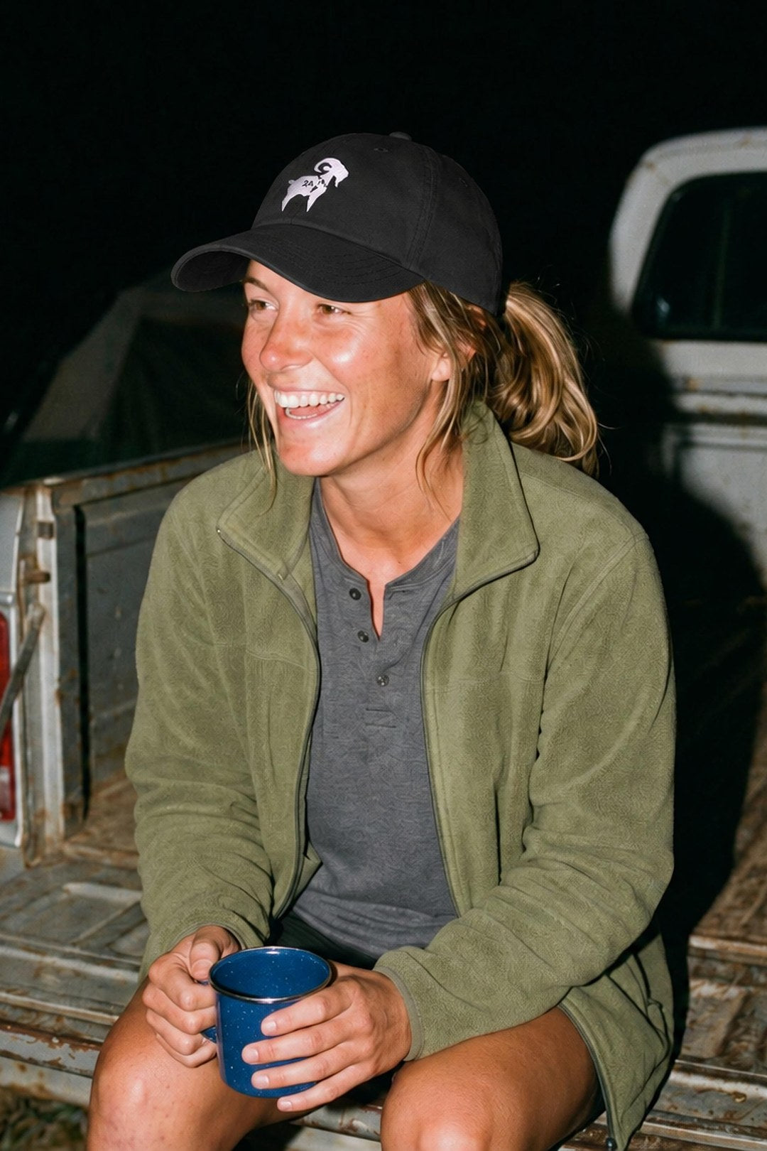 A woman in the black SALTYmf 2A GOAT dad hat laughs on a truck tailgate at deer camp