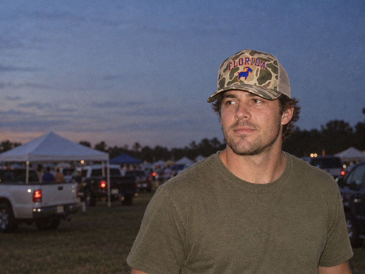 Man in a camo Florida GOAT trucker hat at a tailgate field at dusk