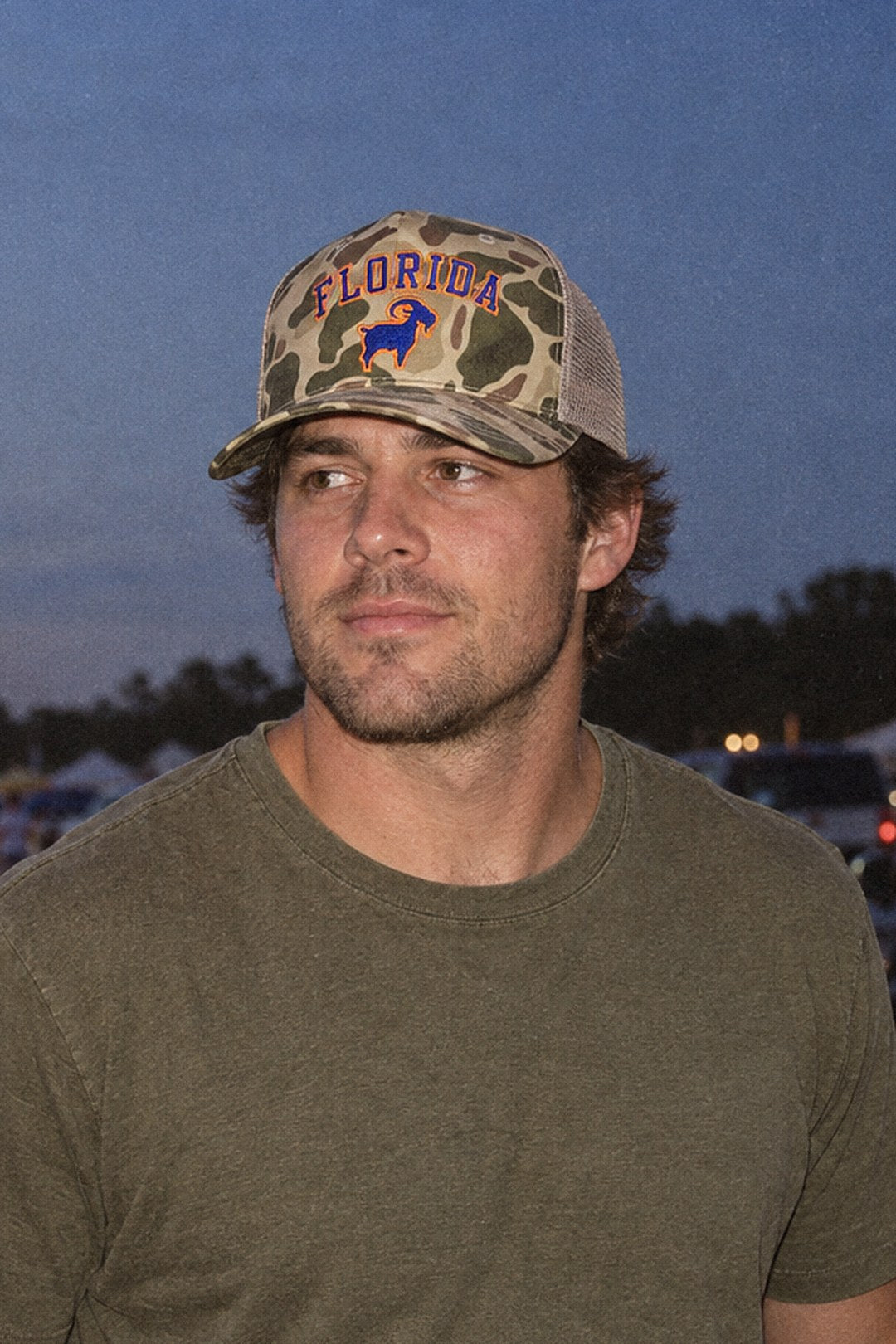 Man in a camo Florida GOAT trucker hat at a tailgate field at dusk