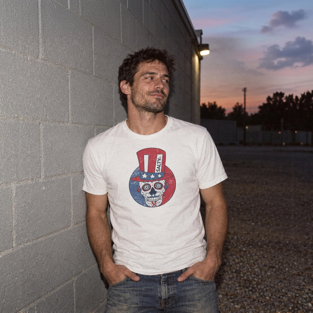 Man in the white SALTYmf Uncle Salty Wants You Tee by a cinder block wall at dusk