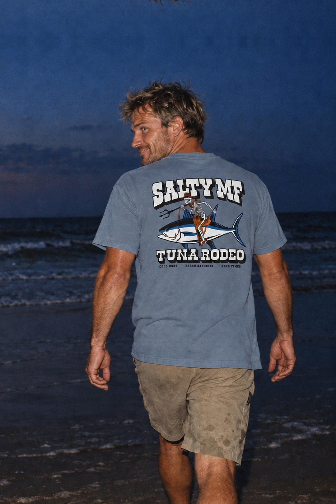 A man in the SALTYmf Tuna Rodeo Tee walks the beach at dusk, the back print facing the camera
