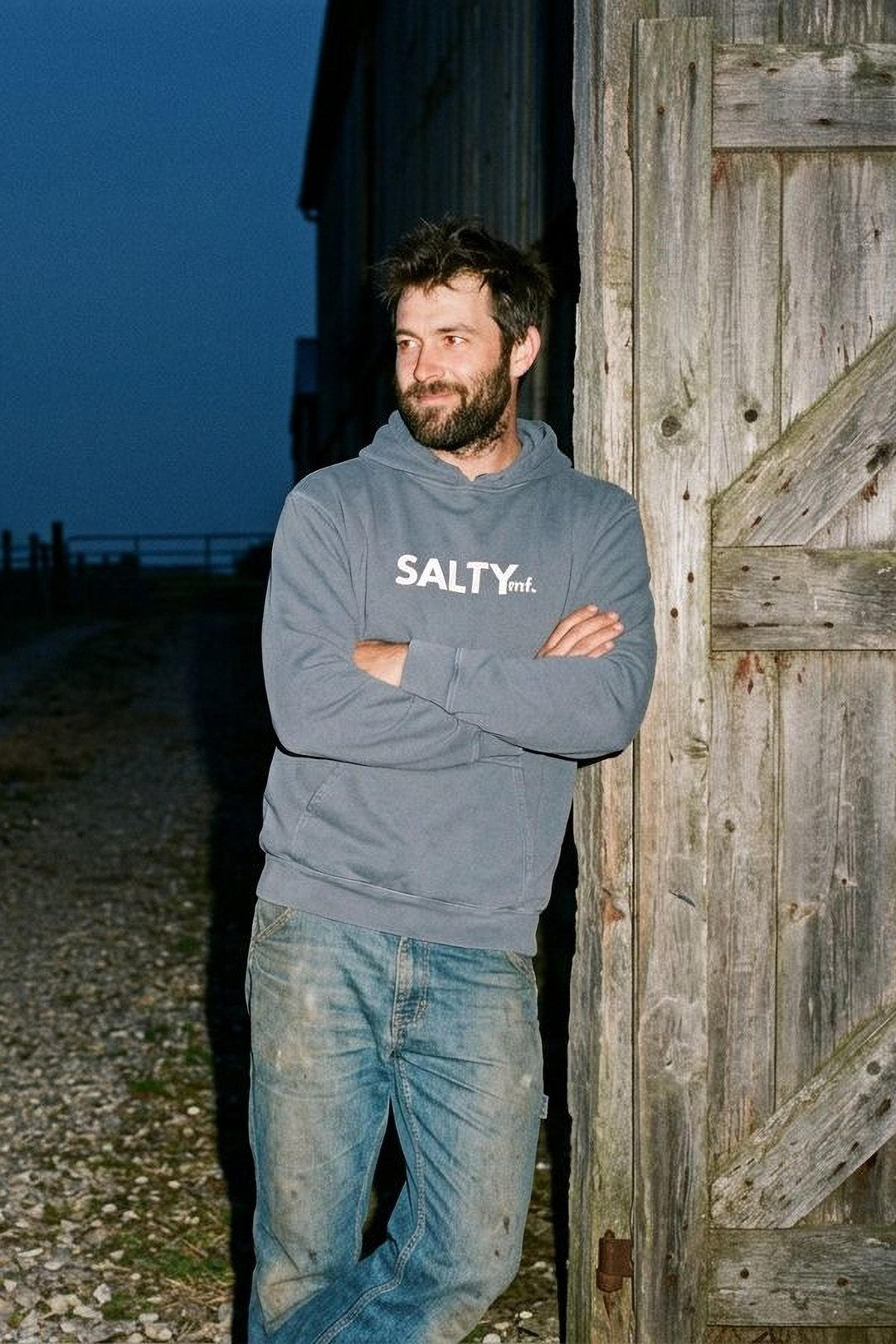 A man in the denim SALTYmf My Salty Hoodie leans on a barn door at dusk